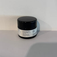 A small black cosmetic jar with a white label, photographed on a light gray surface against a white wall.