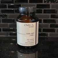 An amber glass bottle of Omica Wellness Shilajit + Amla dietary supplement with a white label and silver cap, set on a dark reflective countertop against a black tiled backsplash.