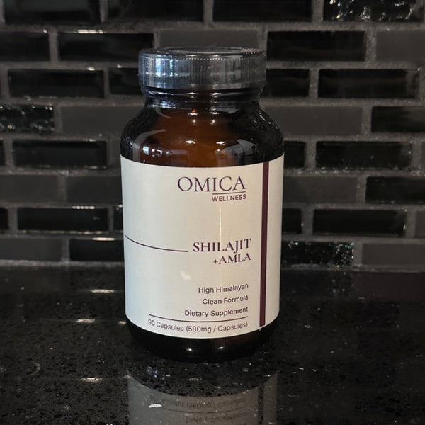 An amber glass bottle of Omica Wellness Shilajit + Amla dietary supplement with a white label and silver cap, set on a dark reflective countertop against a black tiled backsplash.
