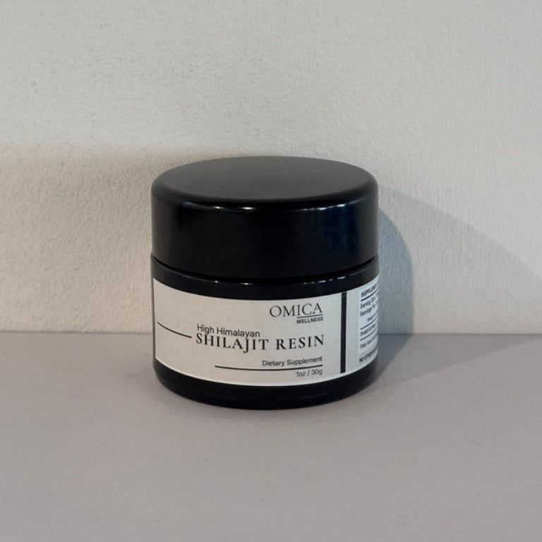 A black plastic jar of Omica Shilajit Resin with a white label, set against a plain light gray wall and surface.