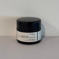 A black plastic jar of Omica Shilajit Resin with a white label, set against a plain light gray wall and surface.