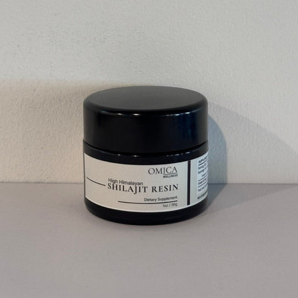 A black plastic jar of Omica Shilajit Resin with a white label, set against a plain light gray wall and surface.