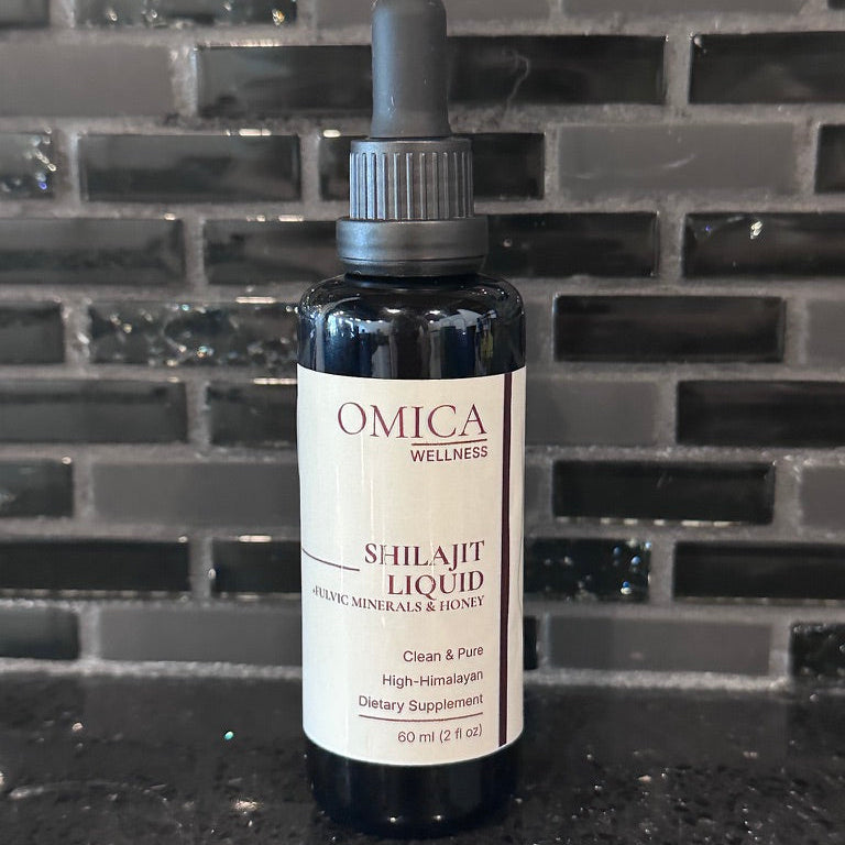 A bottle of Omica Wellness Shilajit Liquid herbal supplement, featuring a dark amber glass bottle, black dropper cap, and a cream-colored label, set against a dark gray tiled background.