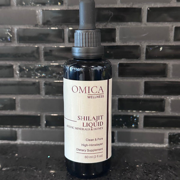 A bottle of Omica Wellness Shilajit Liquid herbal supplement, featuring a dark amber glass bottle, black dropper cap, and a cream-colored label, set against a dark gray tiled background.