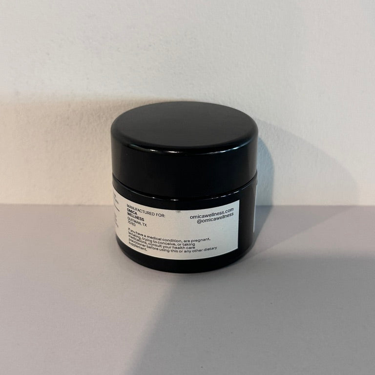 A small black cosmetic jar with a white label, photographed on a light gray surface against a white wall.