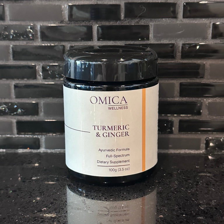 A jar of OMICA WELLNESS Turmeric & Ginger dietary supplement, set on a dark countertop against a dark tiled backsplash.