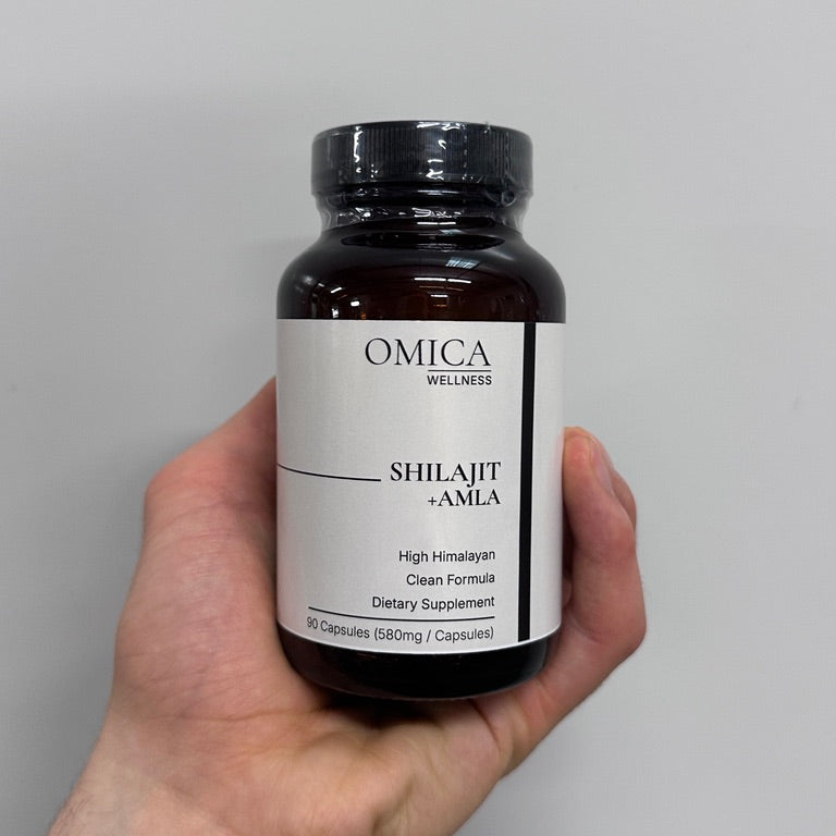 A hand holding a dark amber bottle of OMICA Wellness Shilajit + Amla dietary supplement capsules against a plain light gray background.