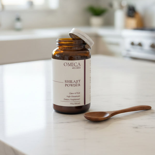 A bottle of Omica Wellness Shilajit Powder dietary supplement, set on a dark countertop with a dark tiled backsplash behind it.